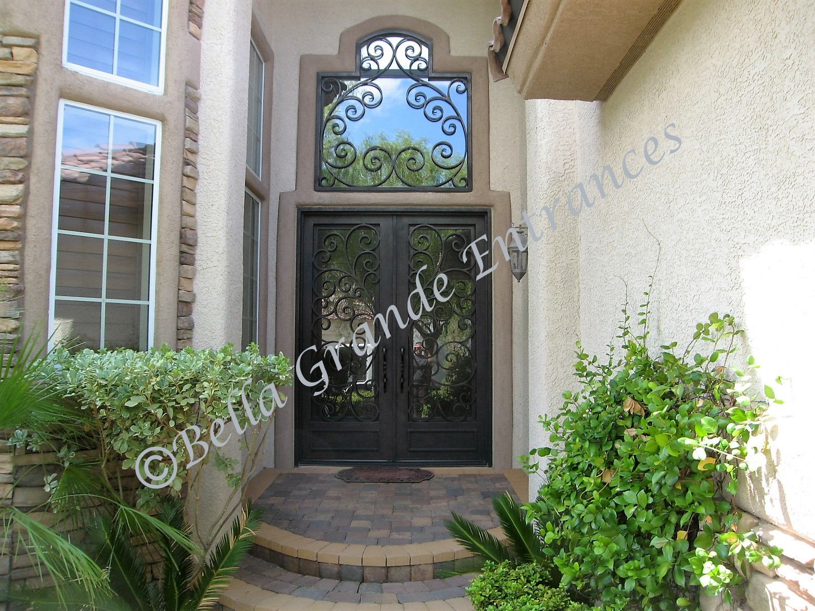 This is a picture of a double ornamental iron door with glass inside. It has a twisted iron handle and a stunning iron grill on top, also made with glass inside. These beautiful doors were proudly crafted by Bella Grande Entrances in Las Vegas.
