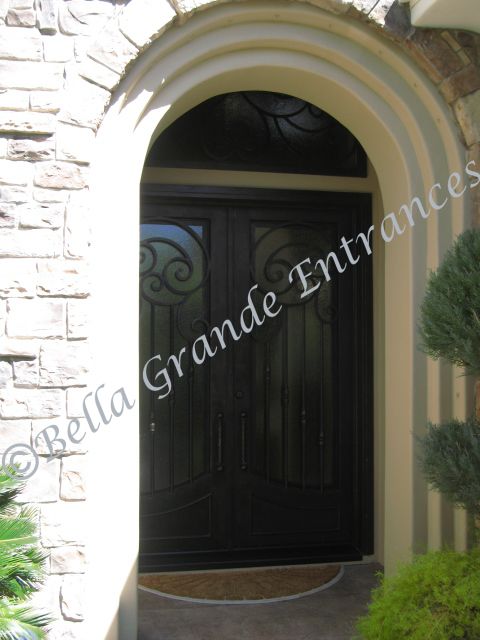 Close-up photo of an elegant iron door. The door includes an iron handle and a window grill at the top. It was proudly crafted by Bella Grande Entrances. This door is installed at a white brick textured exterior house at Bella Grande Entrances.